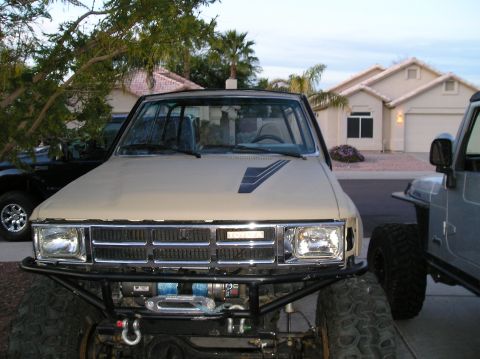 1986 Toyota Hilux - The front view of a tan Toyota pickup truck with a chrome grille and a black brush guard is parked in a driveway. A silver Jeep is parked t…