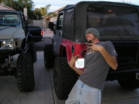 1986 Toyota Hilux - A man wearing a beanie and a grey t-shirt poses with his hands up next to the rear of a red Jeep Wrangler. A lifted Toyota pickup truck is…