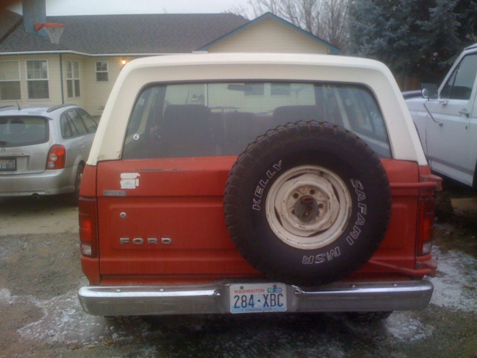 Rear view of a red and white Ford Bronco with a spare tire on the back. Silver hatchback to the left.