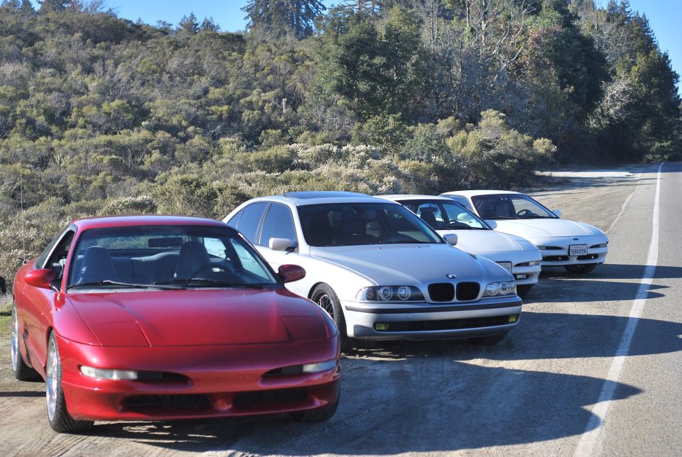 Red Ford Probe parked on a dirt shoulder next to other cars with a wooded hillside.