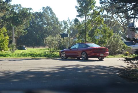 1994 Ford Probe - A red 1994 Ford Probe is parked on an asphalt surface with a grassy area and trees in the background. The car is viewed from the rear-quart…