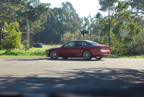 1994 Ford Probe - A red 1994 Ford Probe is parked on the side of a paved road, with trees and a wooden structure in the background.
