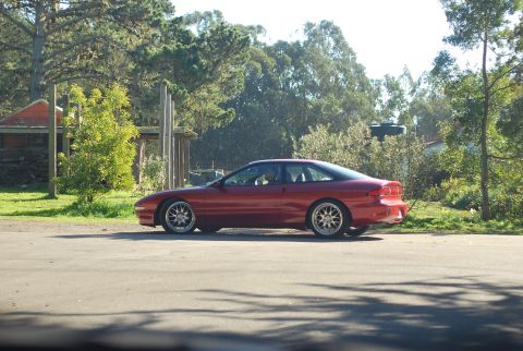 1994 Ford Probe - A red two-door coupe is parked on the side of a paved road. The car is viewed from the side, with a backdrop of trees and a rustic building.
