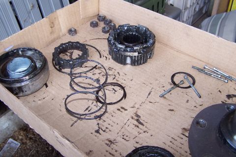 1990 Ford Bronco - A collection of disassembled automotive parts, including gears, nuts, and bolts, are laid out on a greasy cardboard surface within a cardbo…