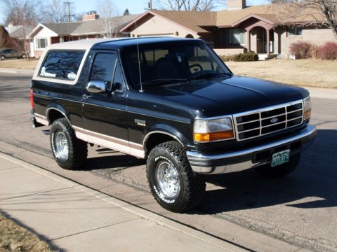 1996 Ford Bronco - A black 1990s Ford Bronco with a tan roof and beige trim sits parked on a residential street. The SUV has chrome accents, off-road tires, a…