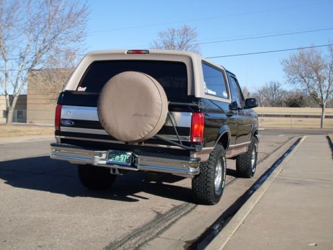 1996 Ford Bronco - The rear drivers side of a black Ford Bronco with a tan top and spare tire cover is parked on asphalt. The vehicle has a chrome bumper and…
