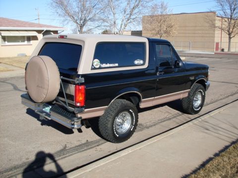 1996 Ford Bronco - The rear driver's side 3/4 view of a black Ford Bronco with a tan top is parked on a street. A spare tire is mounted on the rear door.