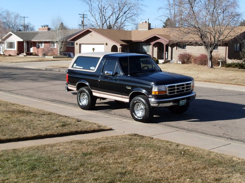 Black 1990s Ford Bronco with tan top parked on street, three-quarter front view.