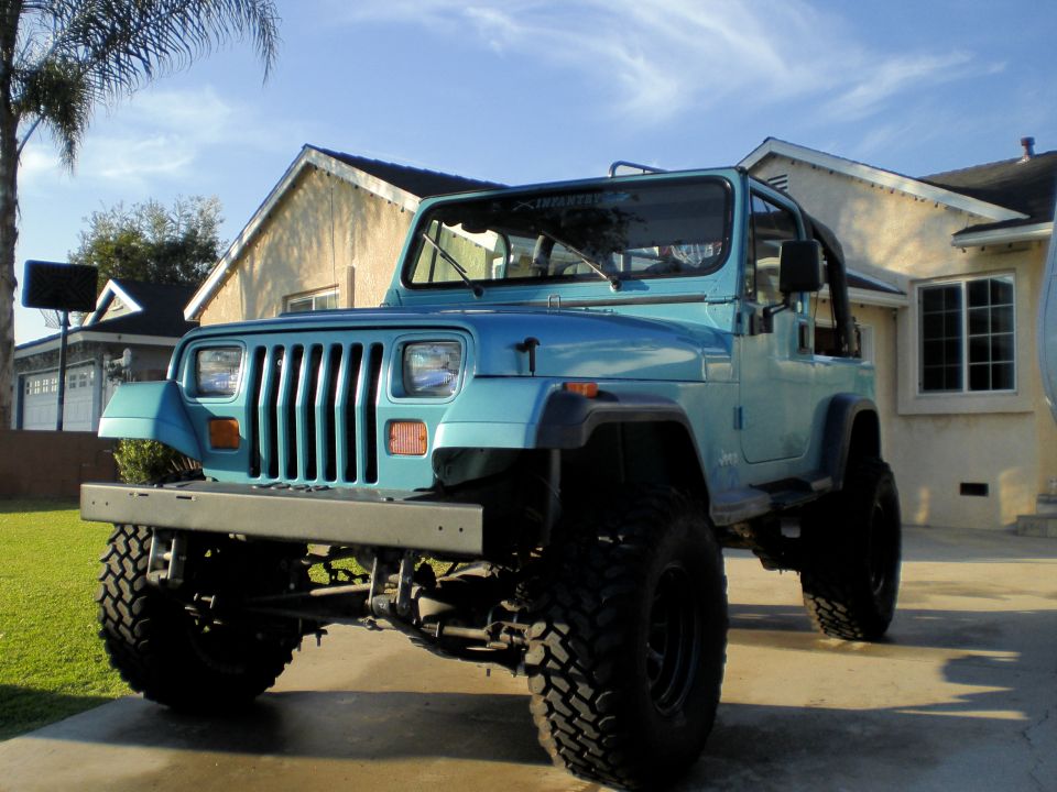 Blue Jeep Wrangler with large tires and aftermarket bumper parked in driveway.