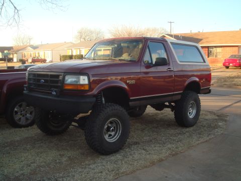 1992 Ford Bronco - A lifted, dark red 1992 Ford Bronco is parked on a patch of grass in front of a suburban house. The vehicle has large off-road tires and a…