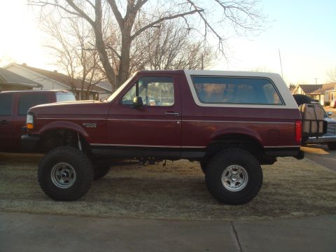 1992 Ford Bronco - The side profile of a lifted, maroon 1992 Ford Bronco with a white top is shown parked on a grassy area in front of houses. It has large of…