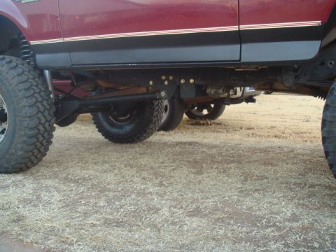 1992 Ford Bronco - The undercarriage of a lifted maroon Ford Bronco is shown, featuring large off-road tires and suspension components. The vehicle is parked…