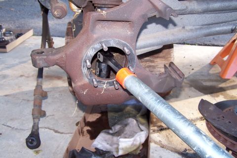 1990 Ford Bronco - A close-up view of a rusty automotive steering knuckle assembly with a socket wrench and extension being used to turn a bolt. A brake rotor…