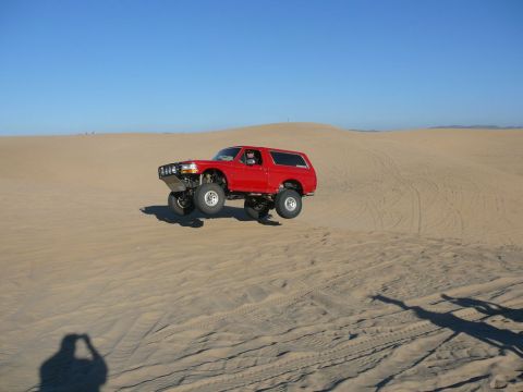 1996 Ford Bronco - A red Ford Bronco is captured mid-air as it jumps over a sand dune under a clear blue sky. The off-road vehicle features a lifted suspensio…