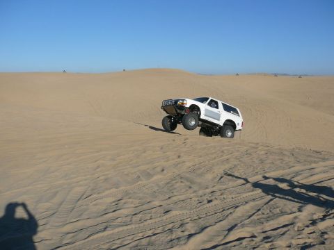 1996 Ford Bronco - A white Ford Bronco is captured mid-air as it jumps over a sand dune in a desert landscape. The vehicle is heavily modified with large tire…