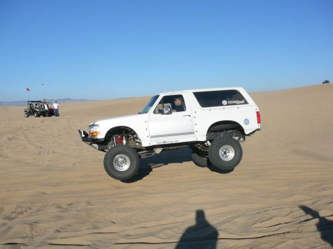 1996 Ford Bronco - A white, lifted Ford Bronco is captured mid-air as it jumps over a sand dune. The vehicle is modified with large off-road tires and suspens…