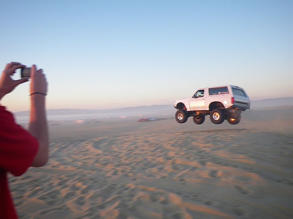 White Ford Bronco jumping over sand dunes, photographed by a person with a phone.
