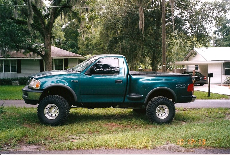 Lifted teal Ford F-150 pickup truck with off-road tires parked on grass.