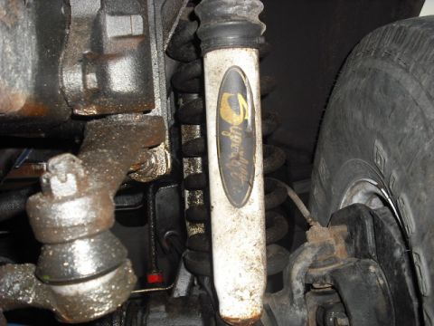 1979 Ford Bronco - A close-up view of the suspension components of a vehicle, including a dirty white shock absorber with a gold logo and a tire visible on th…