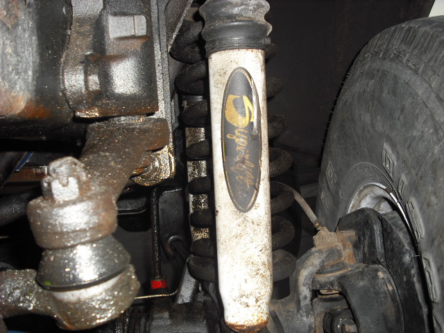 Close-up of vehicle suspension: dirty white shock absorber with gold logo, tire, and steering linkage.