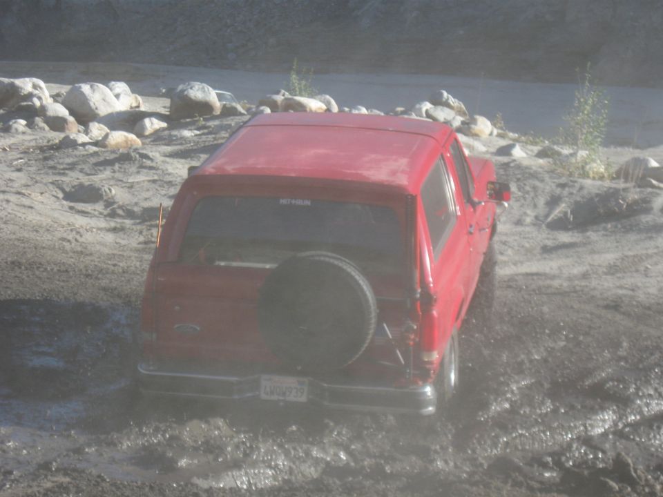 Red Ford Bronco with rear spare tire driving through a muddy rocky wash
