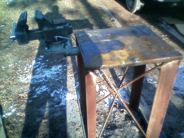 Steel work stand with a black bench vise on a rusty top, set outdoors on gravel.