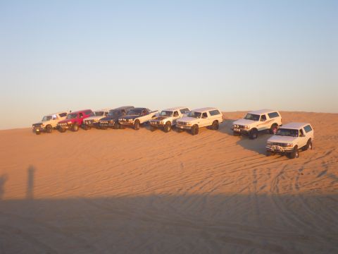 1994 Ford Bronco - A group of Ford Broncos and other SUVs are parked on a sand dune at sunset.