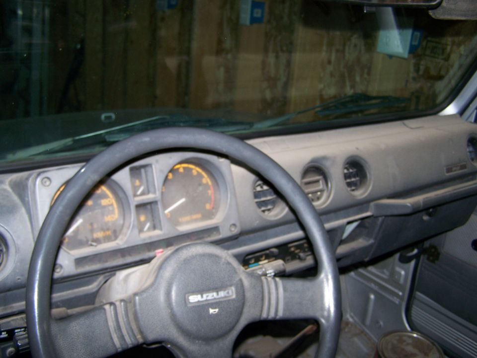 Suzuki Samurai dashboard with steering wheel, gauges, and air vents. "SUZUKI" logo visible on steering wheel
