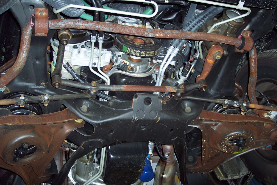 Underside view of a vehicle's engine and suspension components, showing rust on the sway bar and control arms