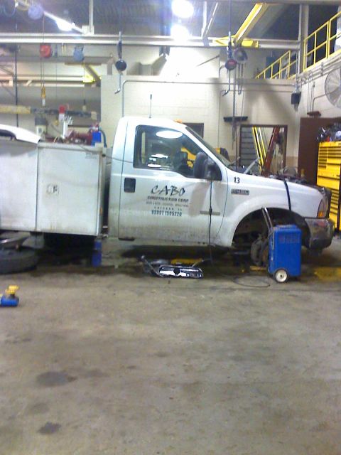 1992 Ford Bronco - A white service truck with a utility bed is undergoing maintenance in a workshop, with its front wheels removed.