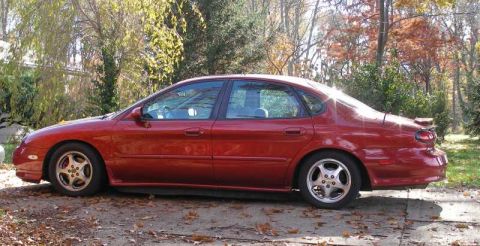 1997 Ford Taurus - A red 1997 Ford Taurus sedan is viewed from the side, parked on a driveway with fallen leaves.