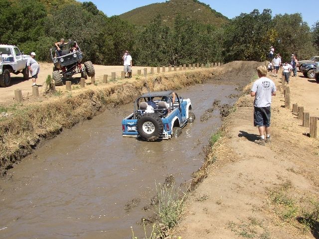 Blue open buggy driving through muddy water trench at off-road park with spectators