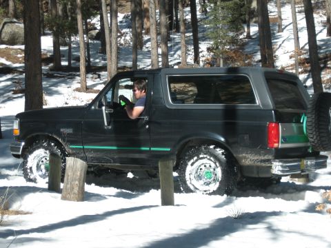 1993 Ford Bronco - A black 1993 Ford Bronco navigates a snowy, wooded area, showing its spare tire mounted on the rear.