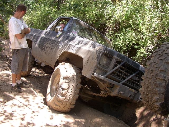 Muddy lifted SUV on large off-road tires climbing rocks on a wooded trail, man watching