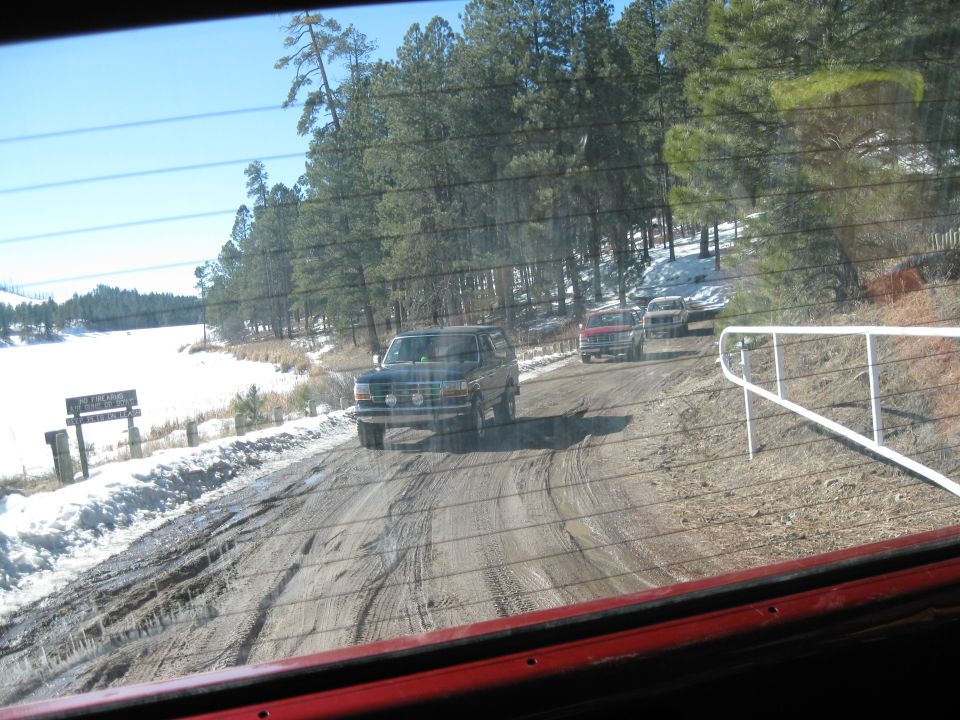 Three SUVs on a muddy, snowy dirt road. The lead vehicle is a dark SUV, followed by a red pickup truck and a white SUV
