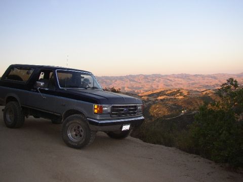 1989 Ford Bronco - This two-tone Ford Bronco is parked on a dirt road with a scenic view of rolling hills in the background.