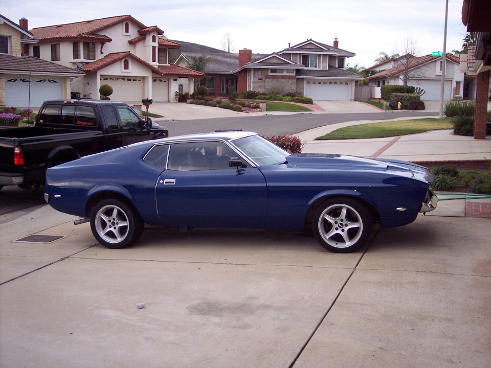 Blue coupe parked in driveway, black pickup truck behind it. Houses in background