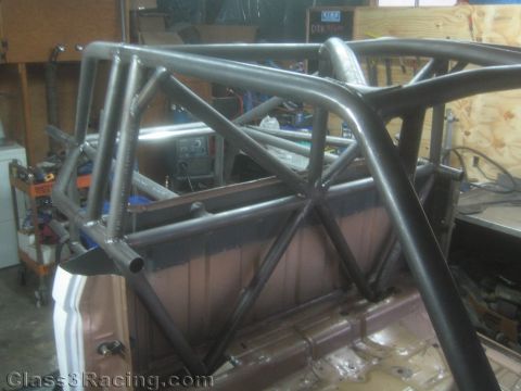 1990 Ford Bronco - Bare-metal and black-painted fabricated roll cage tubing forms a triangulated structure over the rear of a stripped vehicle in a shop.