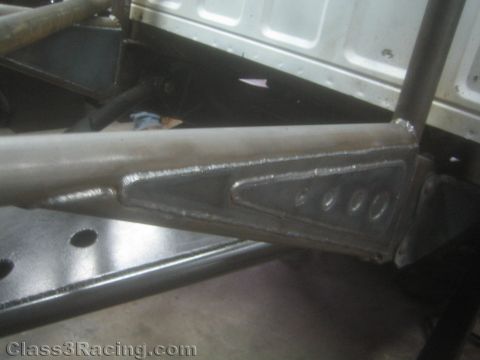 1990 Ford Bronco - fabricated tubular steel suspension arm with a welded, drilled gusset plate resting above a black metal panel in a shop.