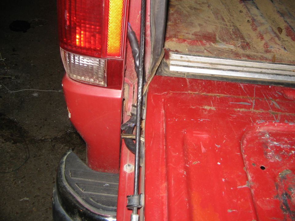 Rear corner of red vehicle showing taillight, chrome bumper, tailgate cable, and worn red cargo bed