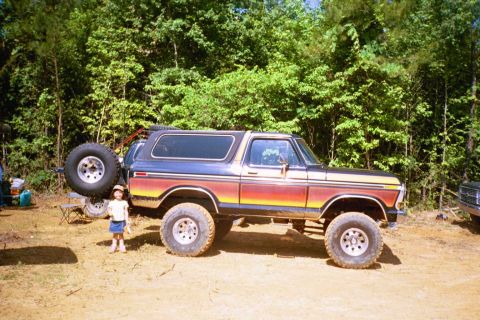 1979 Ford Bronco - A lifted Ford Bronco with a distinctive rainbow stripe paint job is parked outdoors with a child standing nearby.