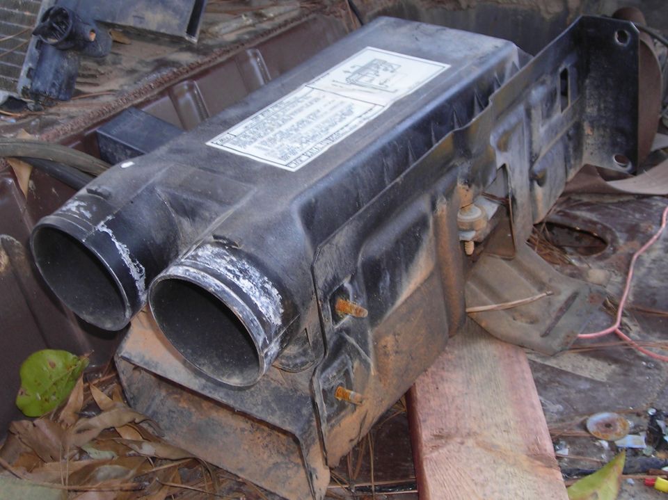 Black plastic vehicle heater blower box with twin round ducts, sitting outdoors on wood and debris