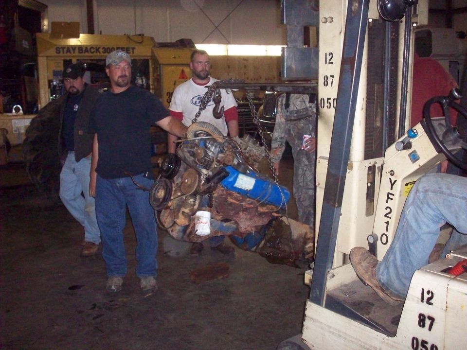 Engine block with blue canister and various hoses being lifted by a forklift with men standing nearby