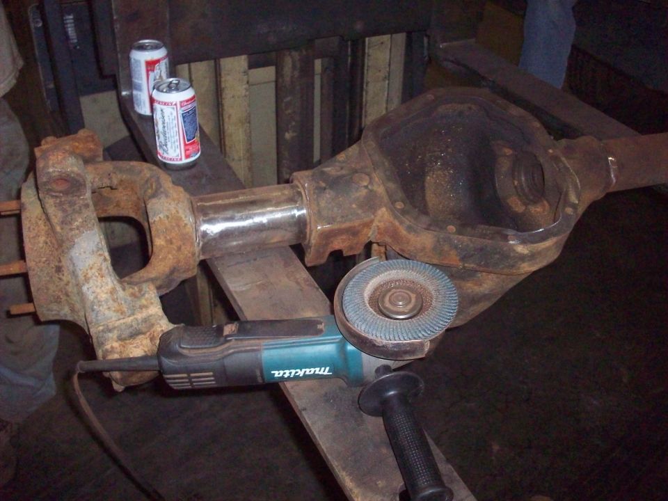 Rusty differential housing and axle assembly on a workbench with a Makita angle grinder and two beer cans