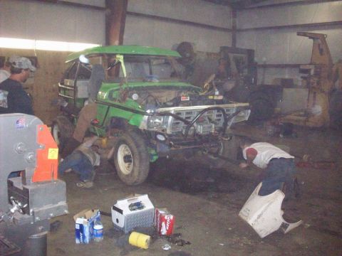 1978 Ford Bronco - Mechanics work on a green off-road vehicle in a garage setting, with various tools and supplies scattered around the floor.