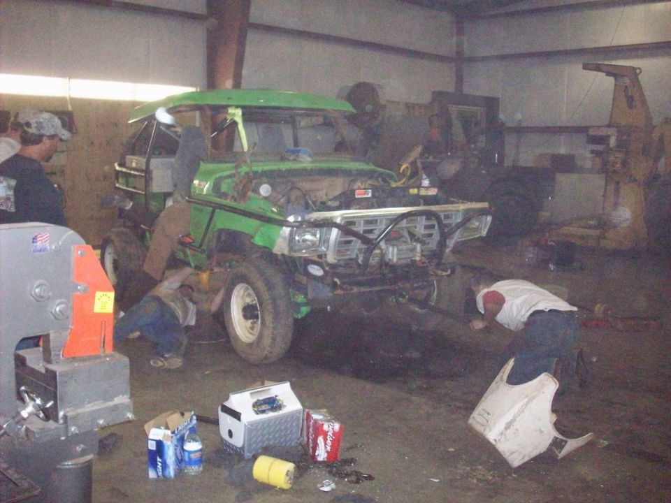 Green off-road vehicle with hood open, being worked on by people in a garage. Some tools and drinks are on the floor