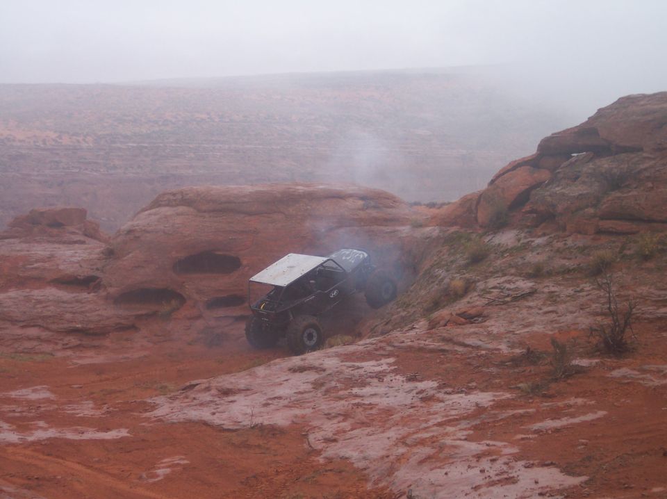 Black off-road vehicle with silver roof climbing a muddy, rocky incline.