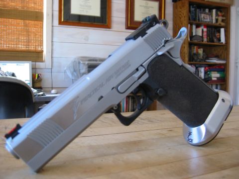 1996 Ford Bronco - A close-up, angled shot of a silver semi-automatic pistol with black grips rests on a wooden table. The pistol has "PRACTICAL ADVANTAGE COM…
