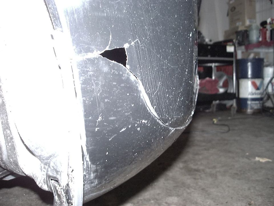 Damaged black car bumper with a large crack and scratches in a garage.