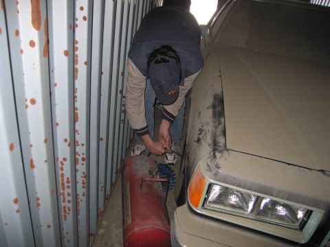 1991 Ford LTD Crown Victoria - Photo of a person working beside a propane tank next to a dusty tan sedan wedged in a storage container.